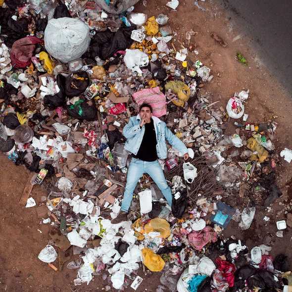 Man making the shush gesture with his finger over his lip, laying on a mound of trash. Photo by Jordan Beltran